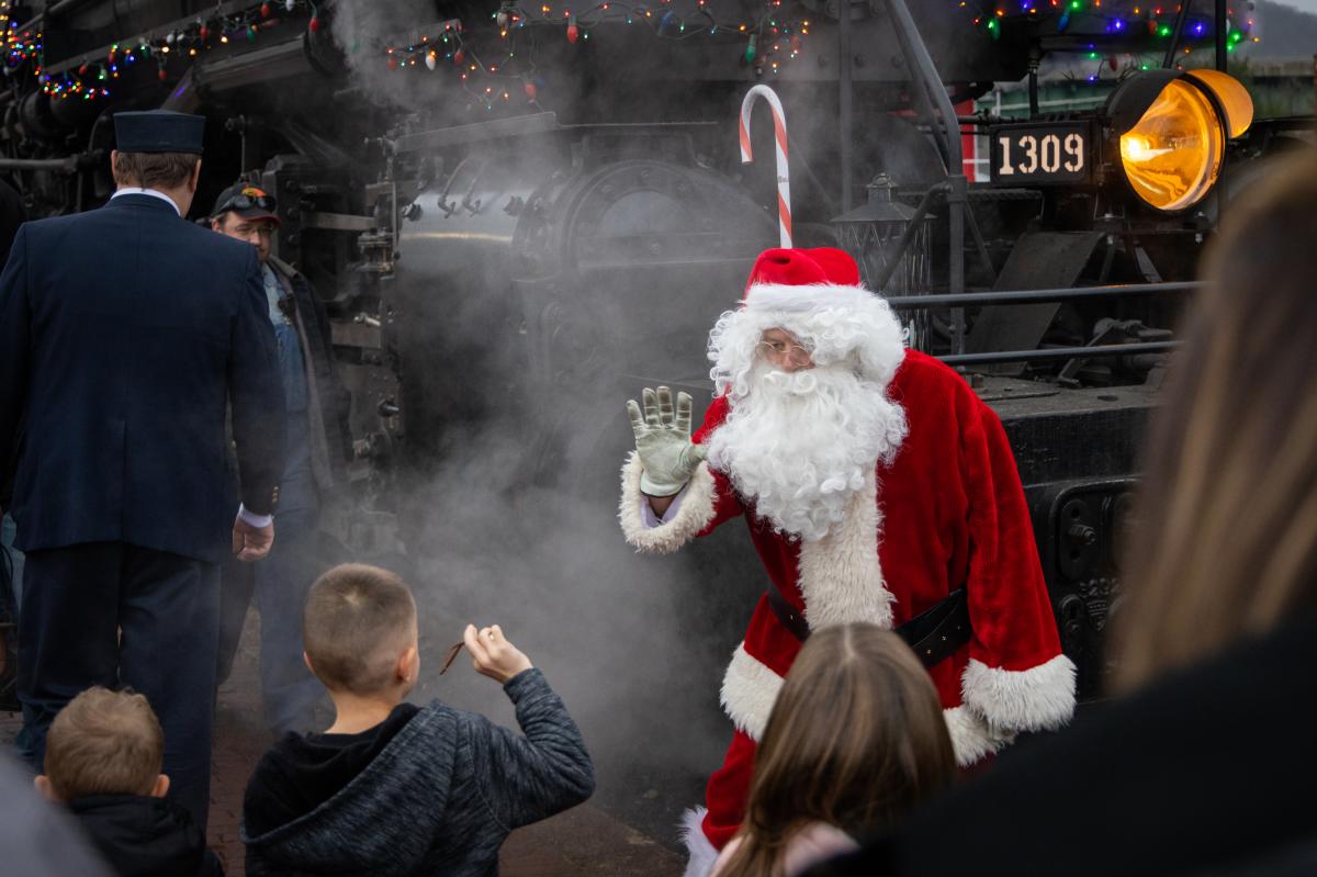 Santa close stands near a steam train engine, waving to a crowd of children.
