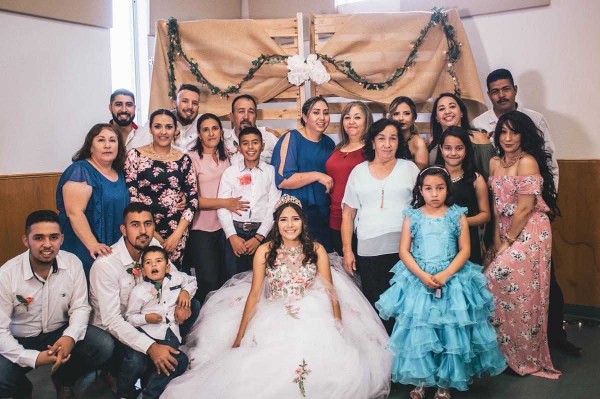 Group photo of hispanic family surrounding the quincenera girl wearing a white dress and crown with flower embellishments