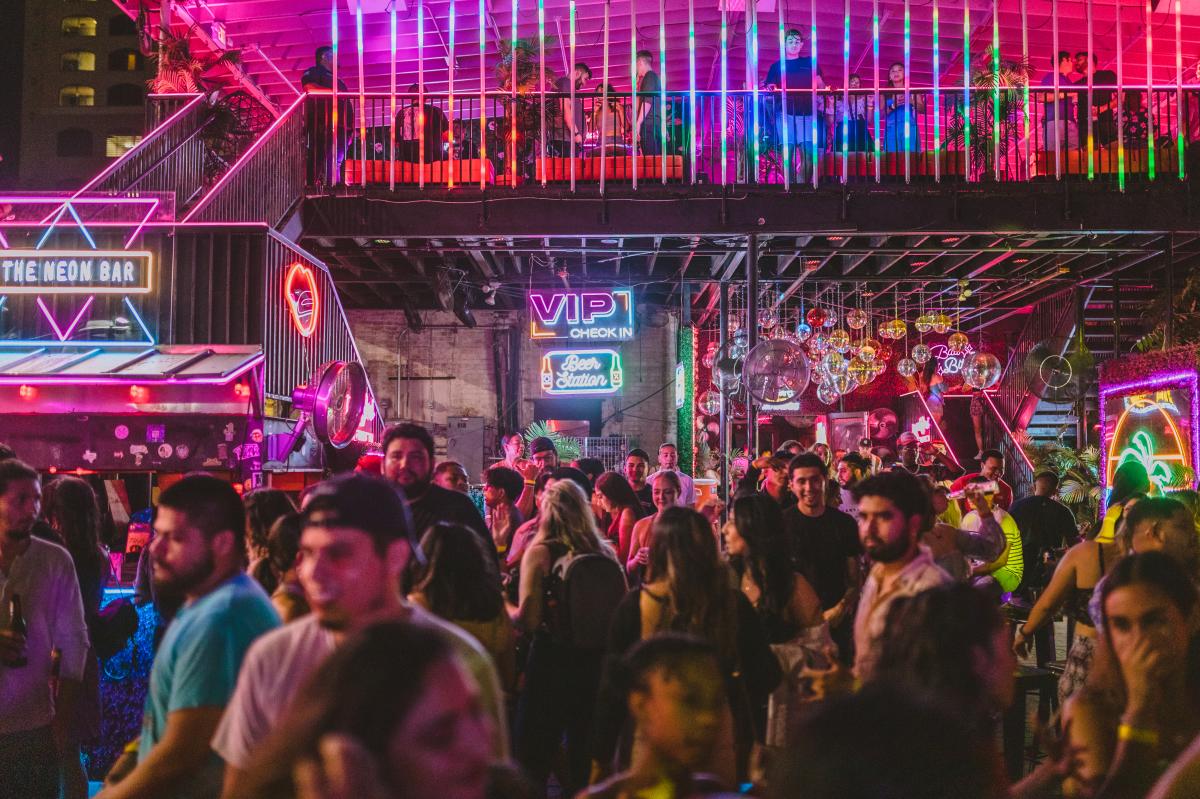 Crowd of people standing on an outdoor patio covered in hot pink, yellow, blue and green neon lights.