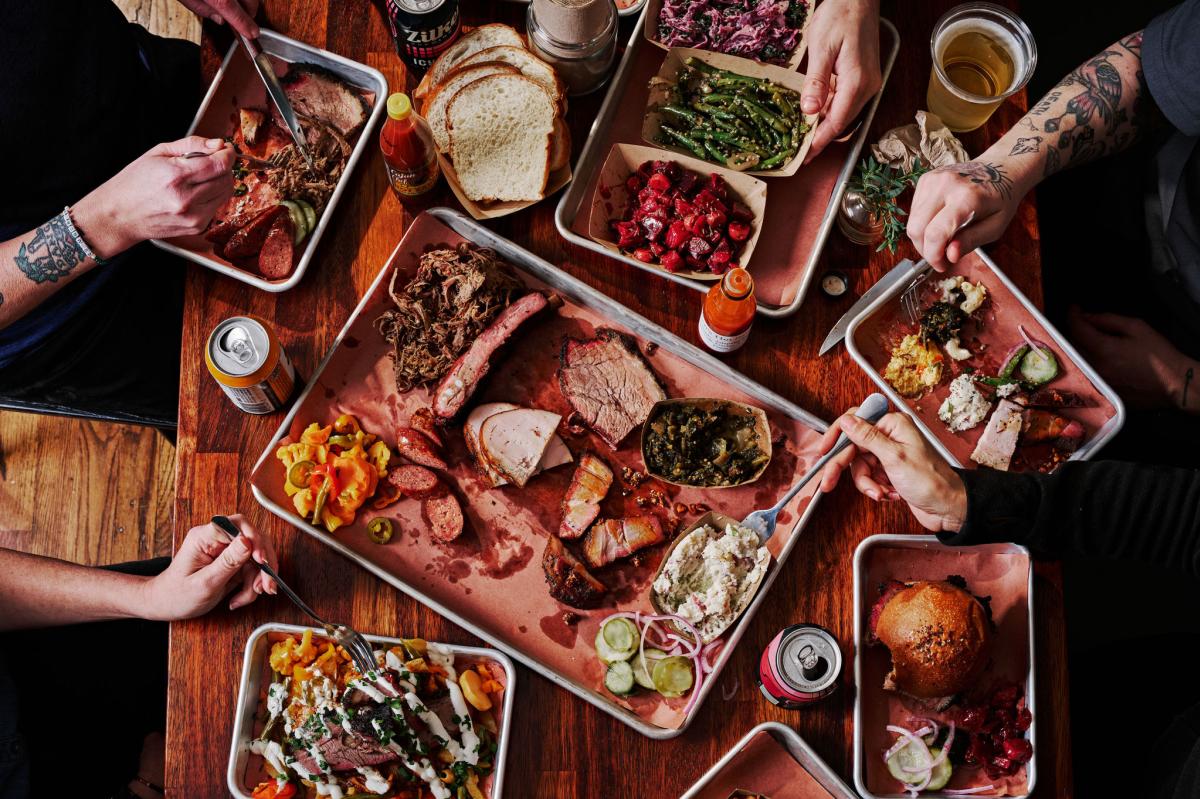 Overhead image of a table full of plates of smoked barbecue and hands reaching into the frame holding utensils and grabbing food.