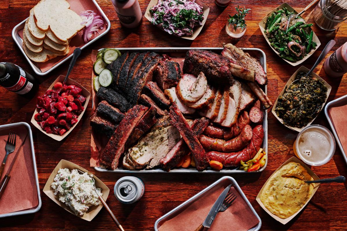 Lay flat image of a table full of a large platter of barbecue meats surrounded by small side dishes and drinks.