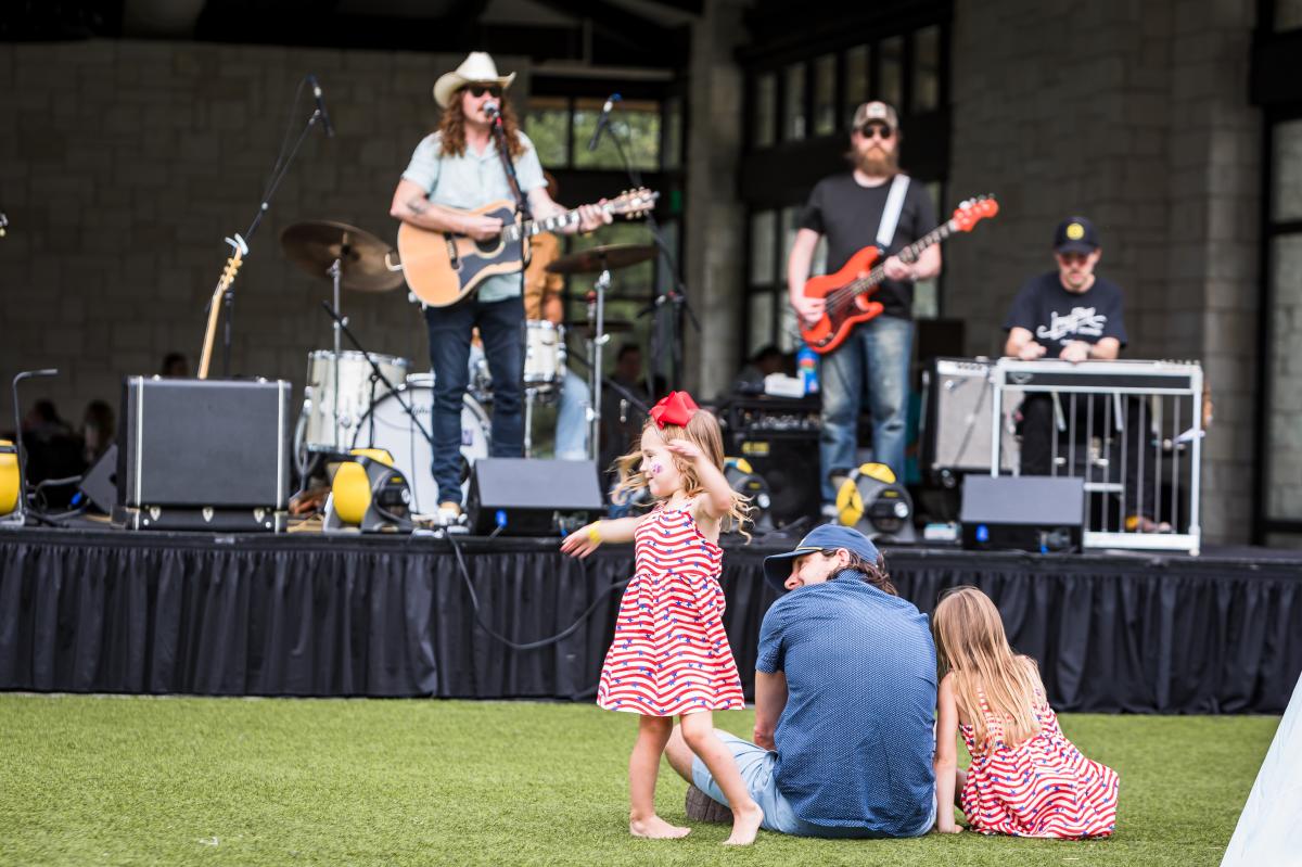 Two young girls and their father sitting together on a grassy lawn listening to live music from the band on the stage in the background.
