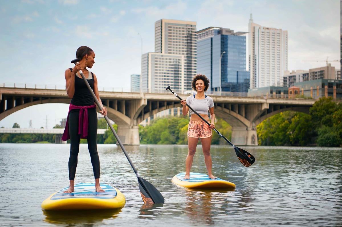 Two women on stand-up paddleboards on Lady Bird Lake with the Congress Avenue Bridge and Downtown Austin in the background.