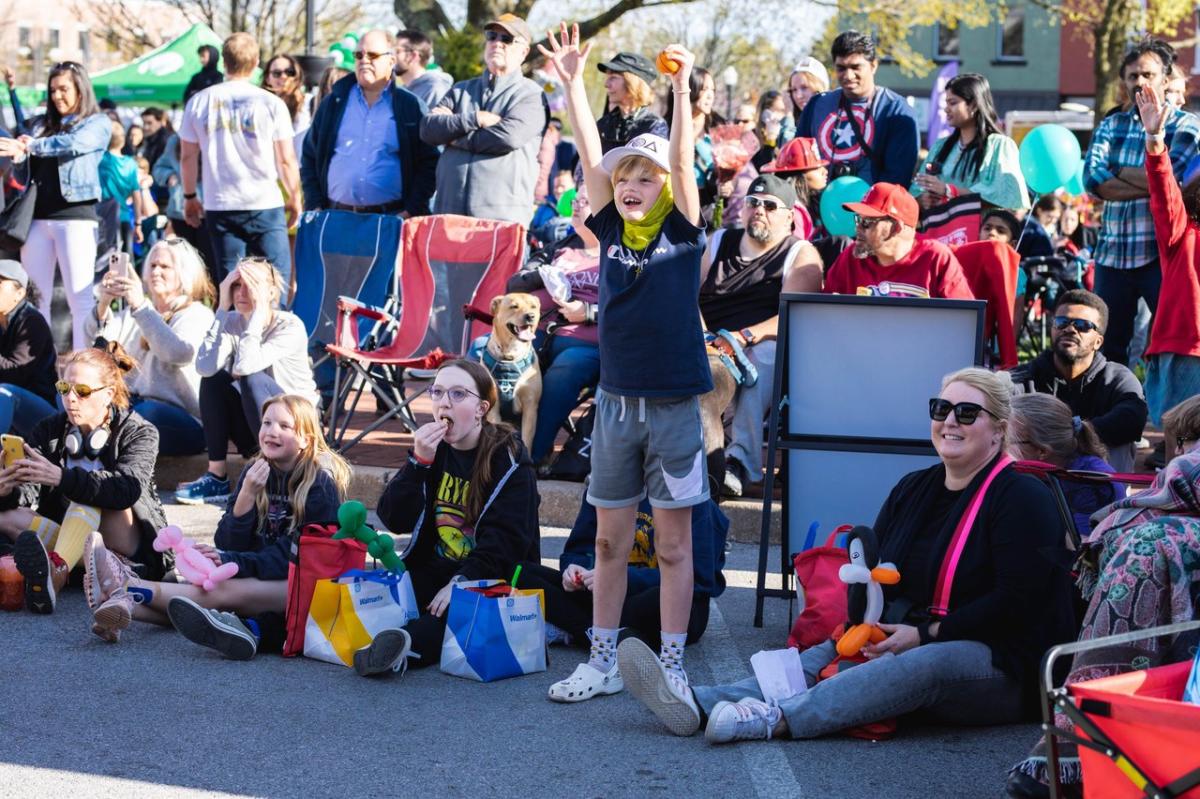 A lively outdoor event with a diverse crowd seated on the ground, including children and adults, enjoying the atmosphere. A boy in a cap raises his arms excitedly.