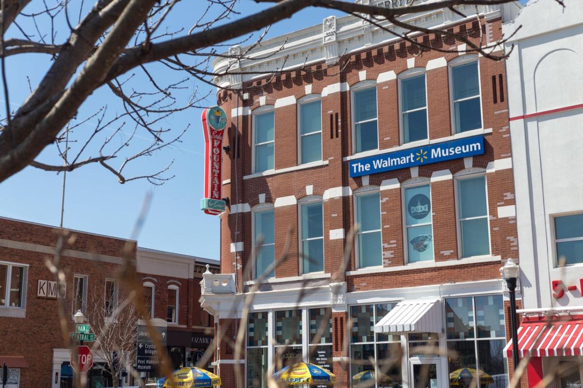 Brick building featuring "The Walmart Museum" sign, with nearby shops and a clear blue sky. A tree branch frames the scene.