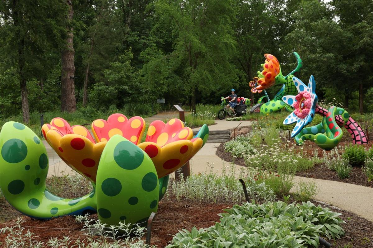 Colorful oversized flower sculptures in a lush garden, with a winding path and greenery. A cyclist rides in the background, enhancing the vibrant scene.
