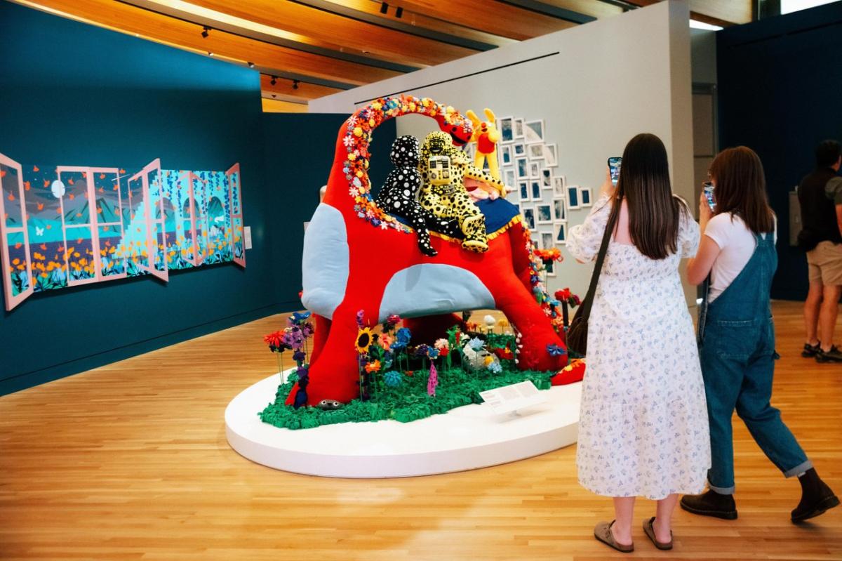 Colorful art installation featuring a large, red sculpture adorned with flowers and a figure, surrounded by visitors in a gallery setting.