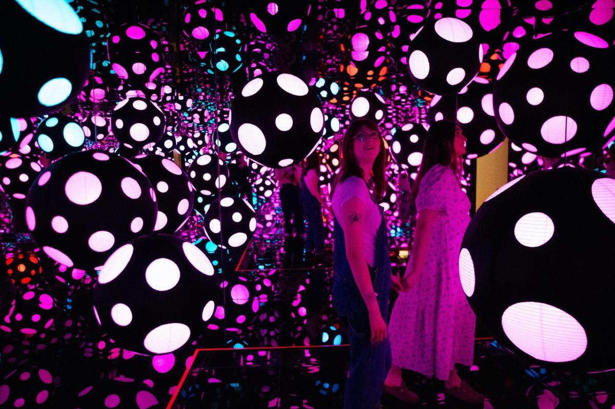 Two visitors explore a vibrant, mirrored room filled with large, illuminated spheres in various colors and patterns. The atmosphere is immersive and dynamic.