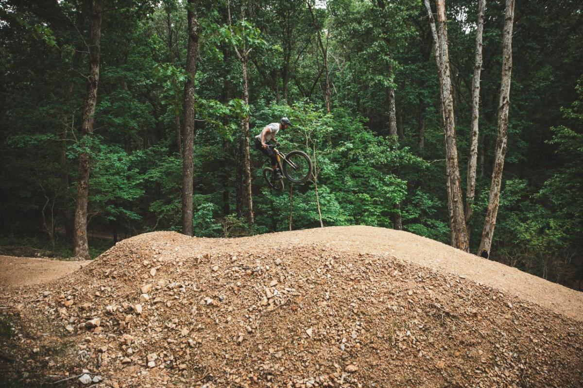 A mountain biker performs a jump over a dirt mound in a lush forest, surrounded by tall trees and greenery. The scene captures an adventurous outdoor activity.