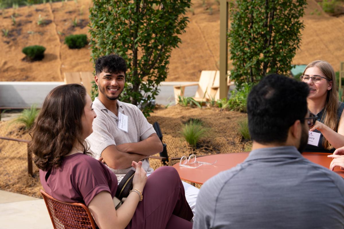 Four people seated around a circular outdoor table in a landscaped garden, engaged in conversation. The table has papers and eyeglasses on it, and one person is wearing a name tag. Faces are blurred for privacy