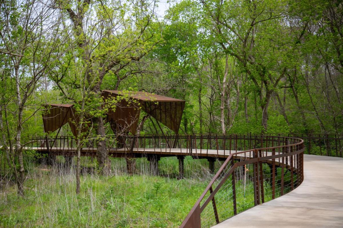 A winding pathway leads to a modern, angular structure nestled among lush green trees, surrounded by tall grasses and nature.