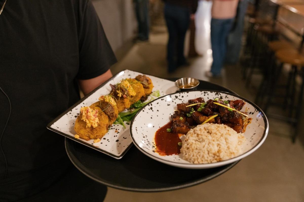 A server holds a black tray featuring two plates: one with golden fritters topped with herbs, and another with glazed meat, rice, and sauce.