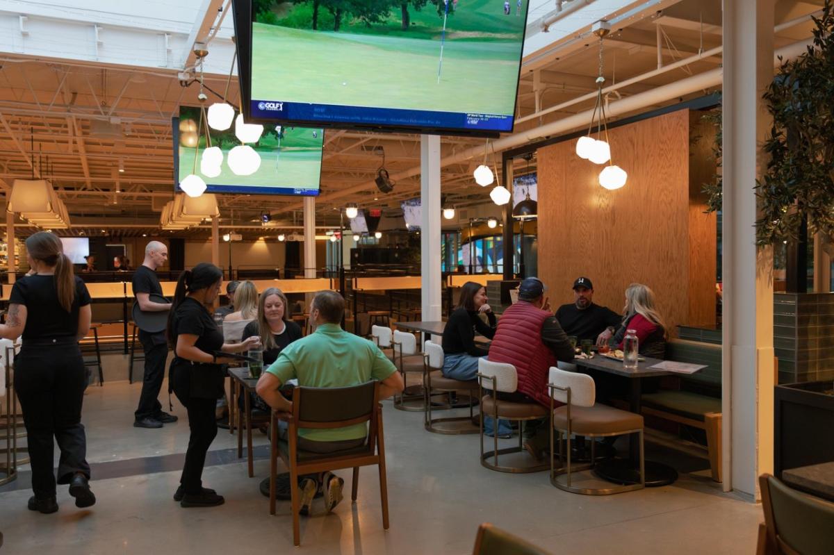 A lively dining space with groups of people enjoying meals, large screens displaying sports, and staff serving at tables. Modern decor enhances the atmosphere.