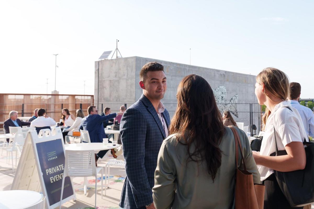 A rooftop venue hosts a private event with guests mingling at tables. A man in a suit engages with two women, surrounded by a cityscape.