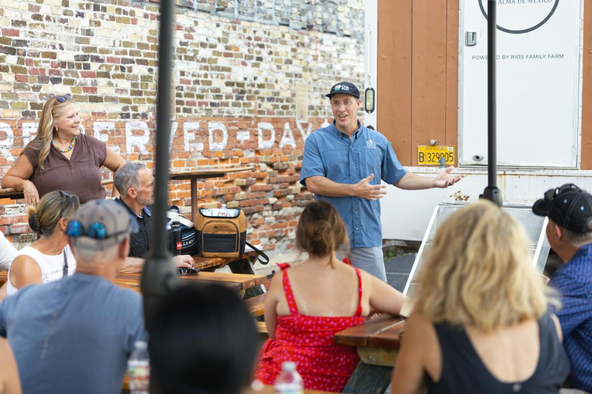 A food tour guide speaks to a group gathered at outdoor picnic tables in downtown Bentonville, Arkansas, with a food truck and brick building in the background.