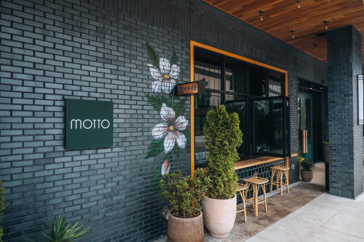 Exterior view of a modern café named Kohi Coffee within the Motto Hotel featuring a floral mural, outdoor seating, and greenery in planters. The inviting entrance showcases a coffee sign.