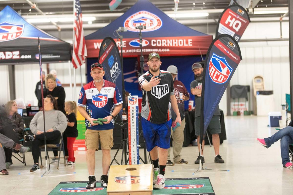 Two male players compete in a cornhole tournament, with colorful banners and spectators in the background. The setting is indoors, showcasing a lively atmosphere.