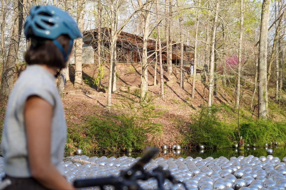 A person wearing a blue helmet and riding a bicycle is partially visible in the foreground, facing a body of water covered in numerous reflective silver spheres. Beyond the water, a wooded area with bare trees and a building on a slight hill are visible under a bright sky.