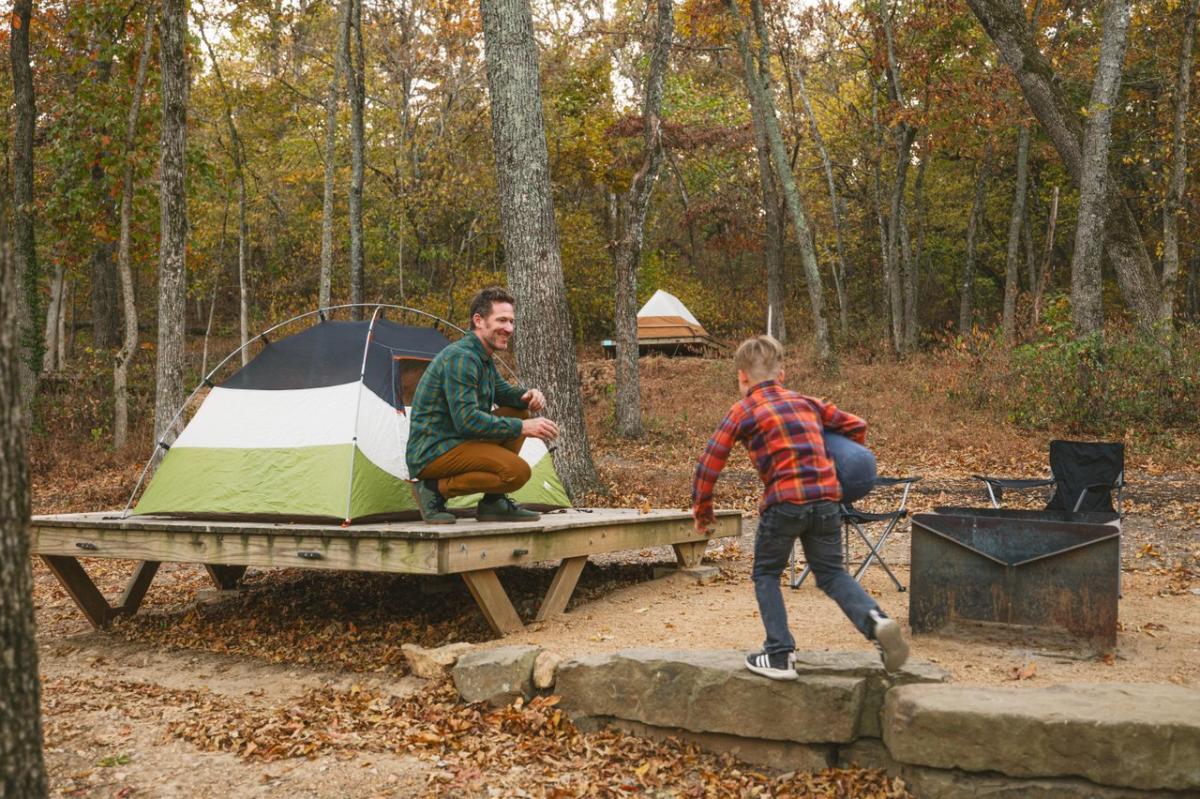 A man sits on a wooden platform next to a tent, smiling at a boy running towards him. Surrounding trees display autumn foliage.