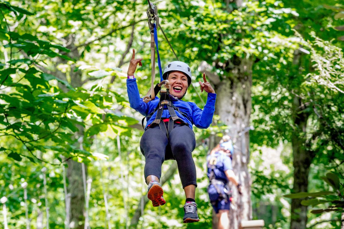 A person wearing blue and gray holds up their hands enthusiastically while harnessed into a zipline.