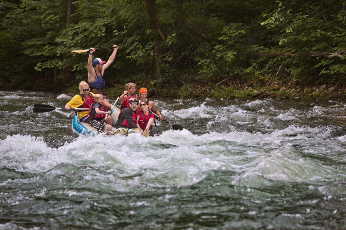 A raft with four rowers and a whitewater rafting guide with his hands raised over his head traverse the rapids of a mountain river.