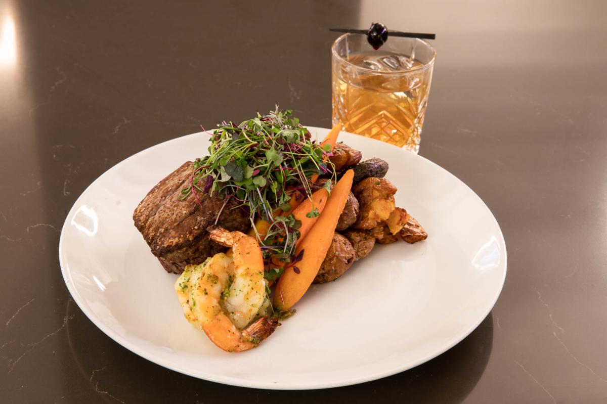 Plated steak with shrimp, carrots, potatoes and a garnishment on top. Cocktail off to the side.