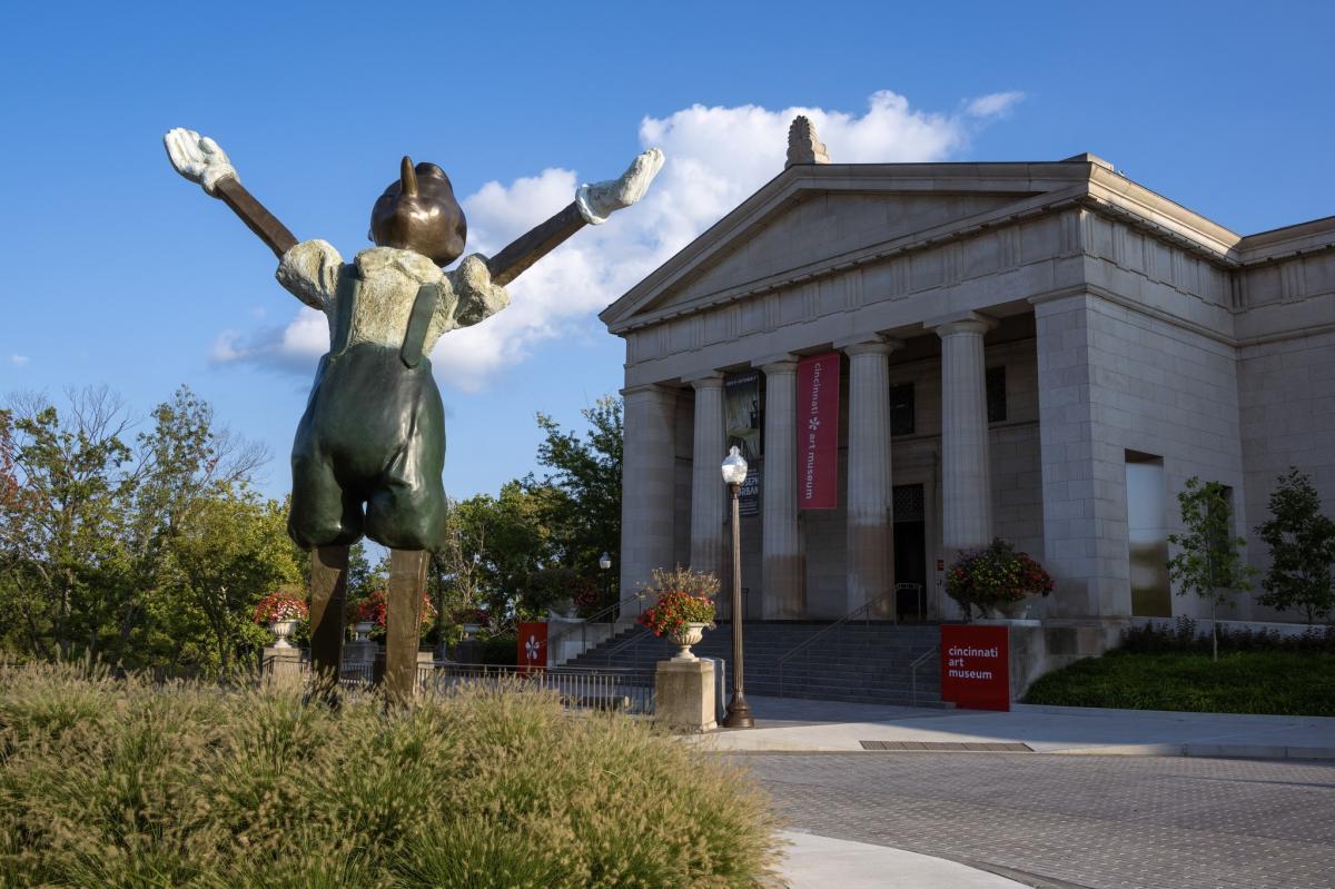 Image is of the Cincinnati Art Museum entrance in the background with a bronze statue of Pinocchio with his hands raised looking to the sky.