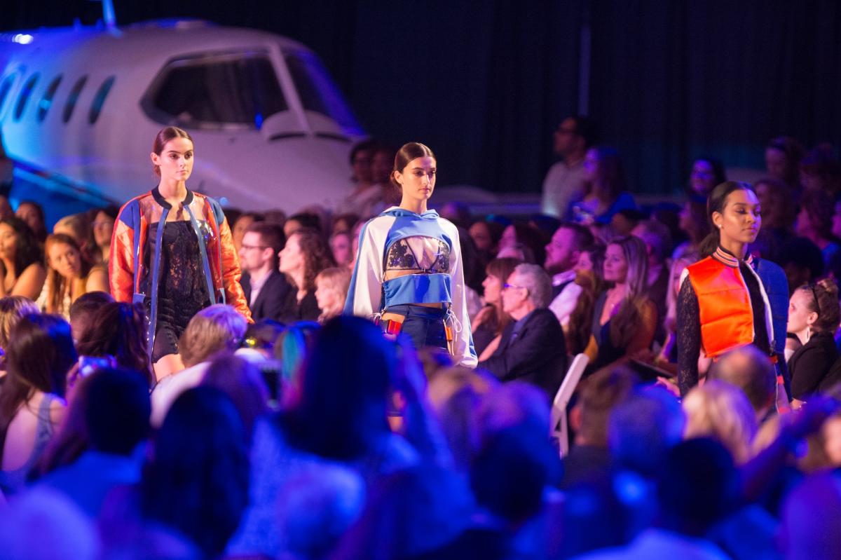 Image is of 3 models walking the catwalk with people and a private plane in the background.