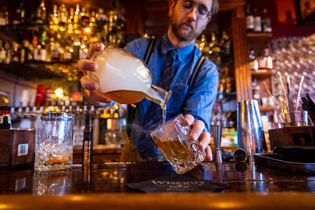 Bartender in blue shirt pouring from a bottle filled with smoke and bourbon