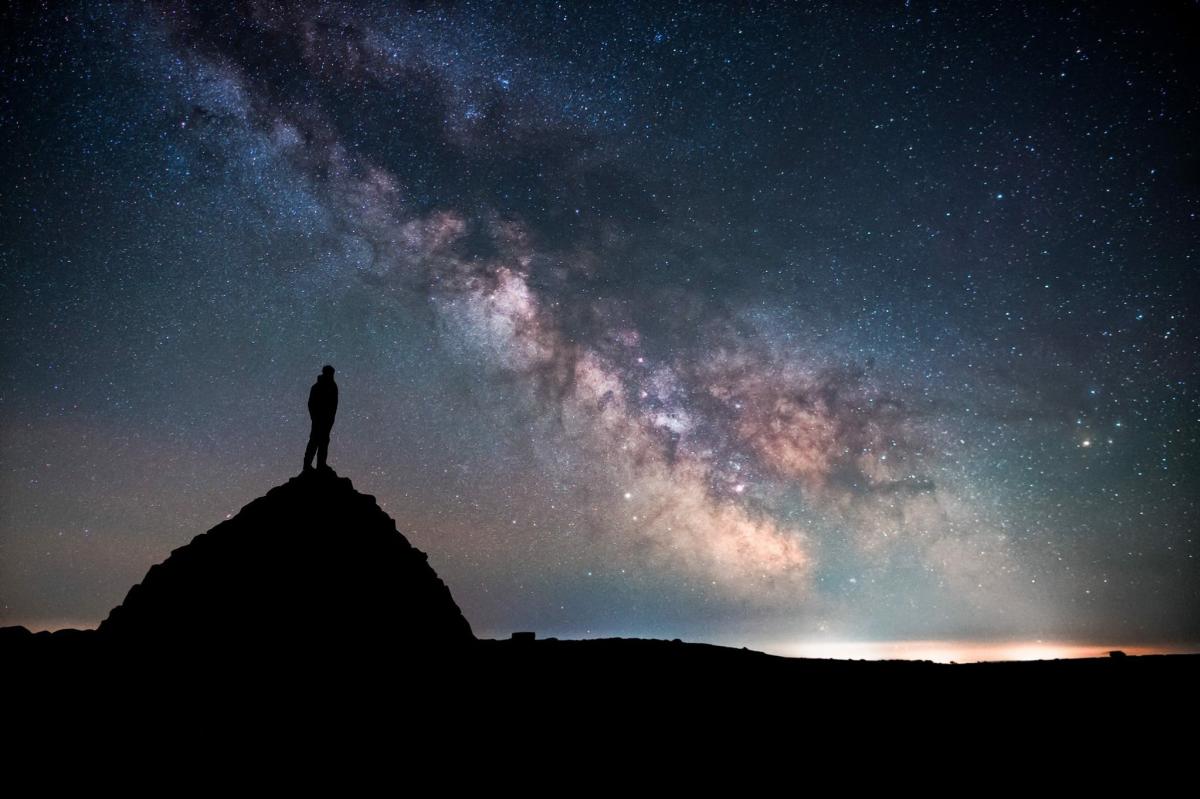 Silhouette of a person standing on a hilltop, gazing at a vibrant, star-filled night sky. The Milky Way arcs across the scene, evoking awe and wonder.