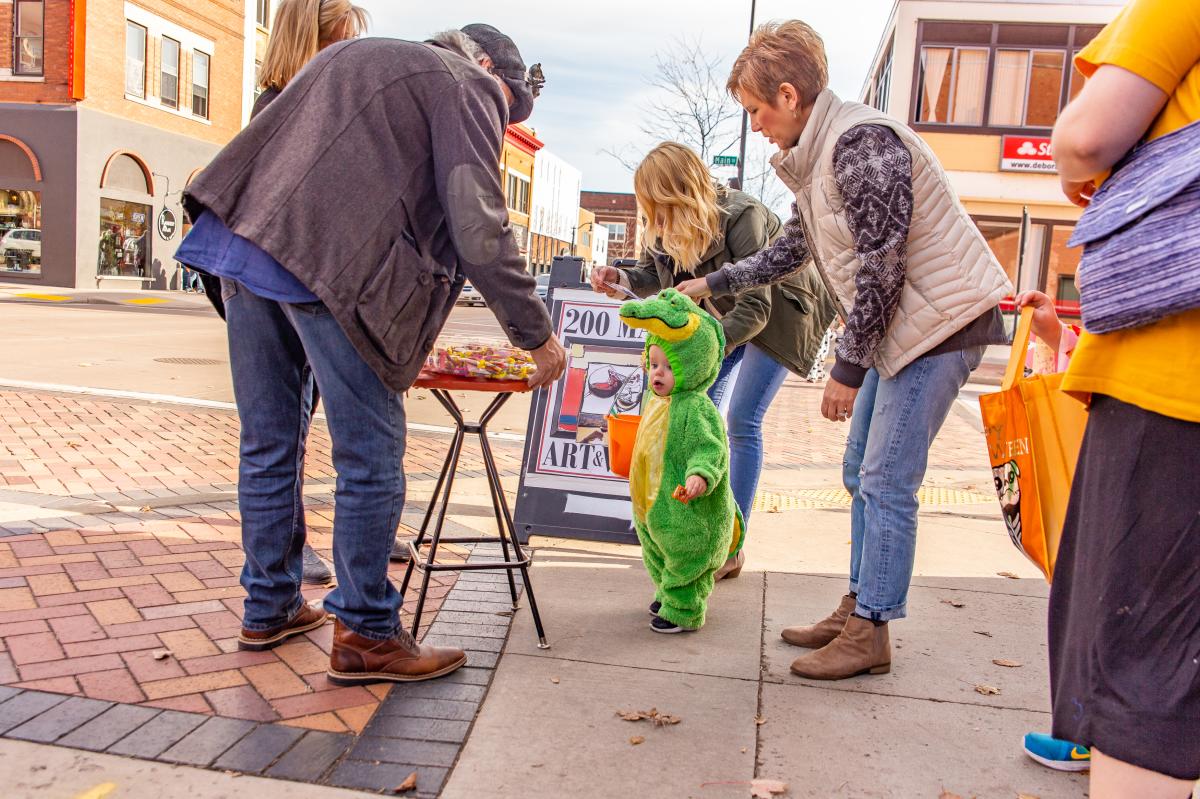 Toddler dressed in a green gator costume trick or treating in Downtown Eau Claire