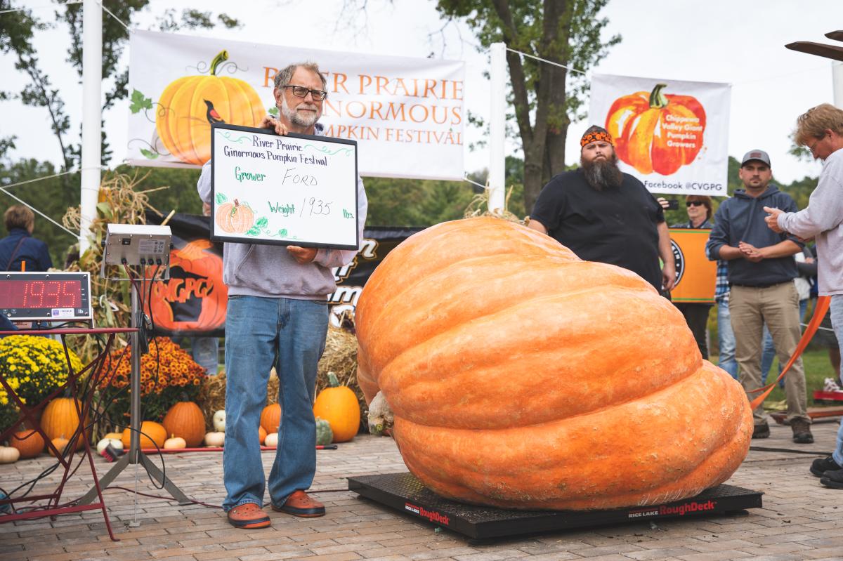 A man posing with a whiteboard sign in front of his giant pumpkin at River Prairie's 2021 Ginormous Pumpkin Festival
