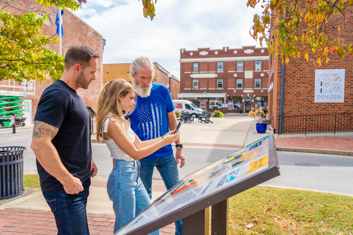 A group of three people, two men and a young woman, stand outdoors interacting with an informational sign in a small town square.