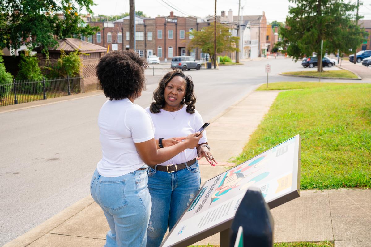 Two women are standing and holding a phone in front of the Kentucky Black Trailblazers plaque for The First Black Baptist Church located in downtown Elizabethtown.