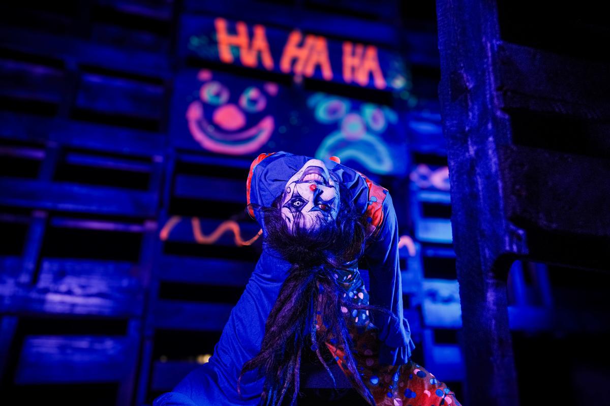 clown-like character bending backwards with a creepy grin in front of a neon background that says "HA HA HA"