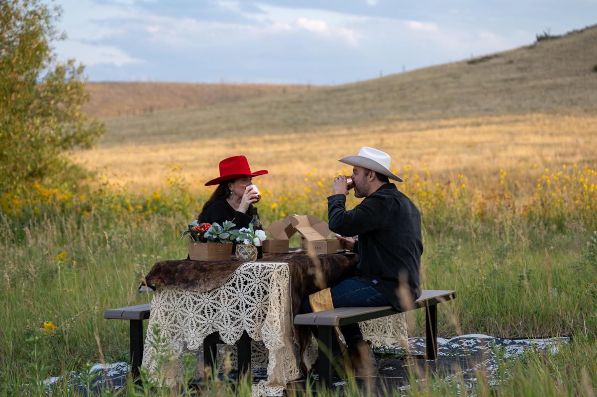 Two people sitting on a picnic table in a field eating charcuterie.