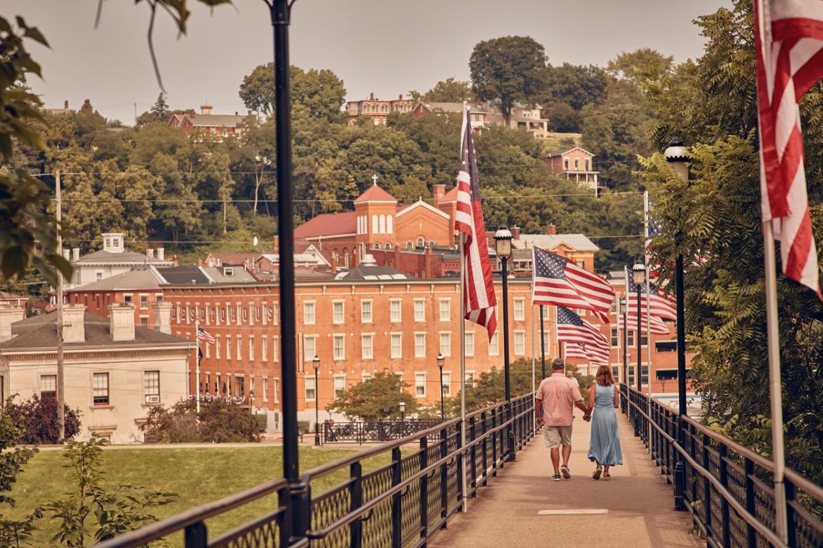 A landscape view of downtown Galena, and the walking bridge, with a couple holding hands walking across it. There are American flags on the bridge, waving in the wind.