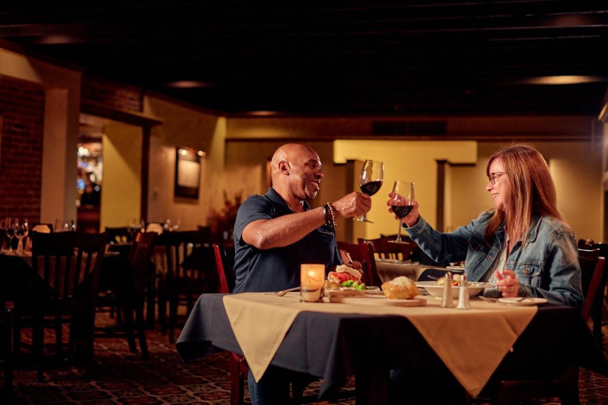 Two people sitting at a dining room table, holding wine glasses, going in for a "cheers".
