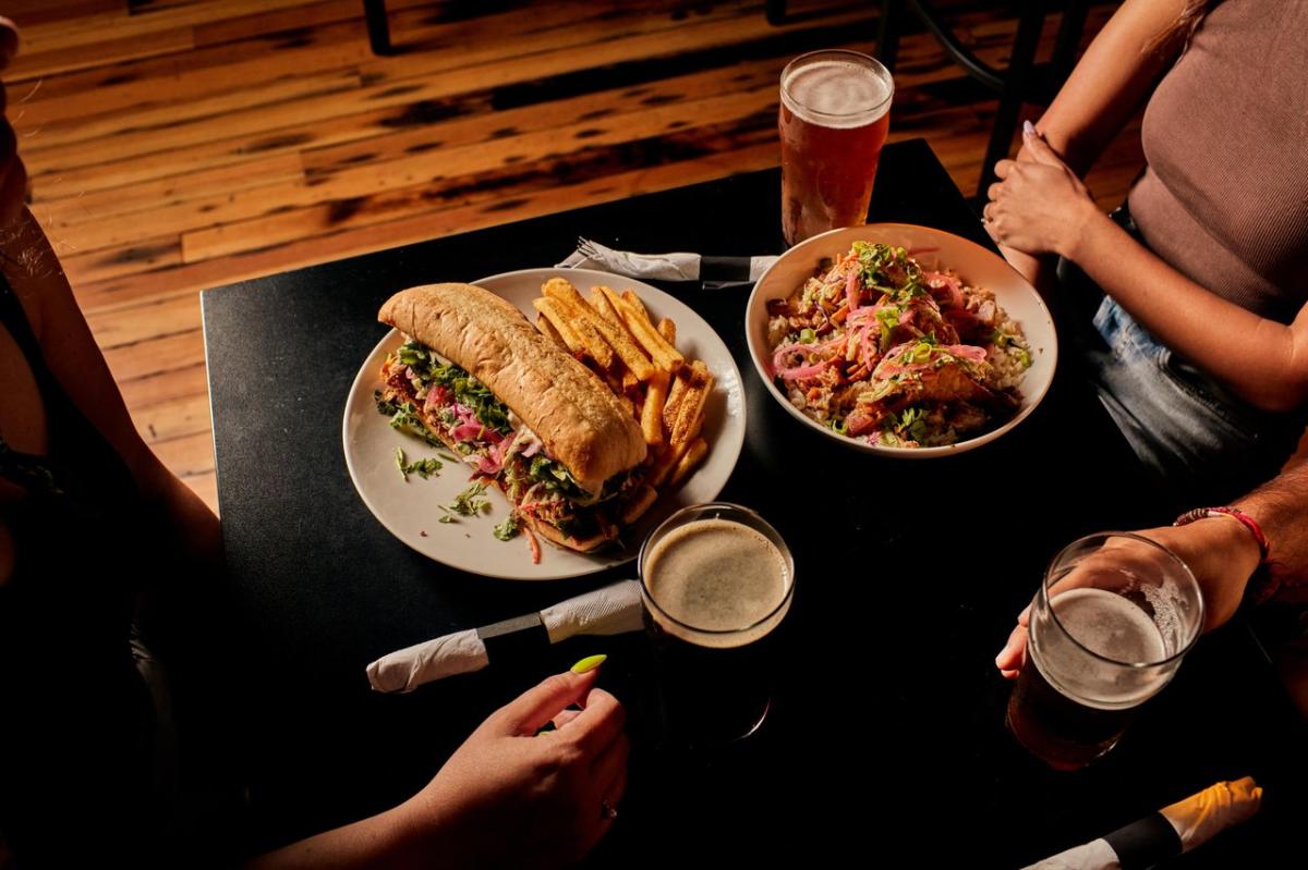 Three people sitting at a table with two plates of food, and beers in glasses.