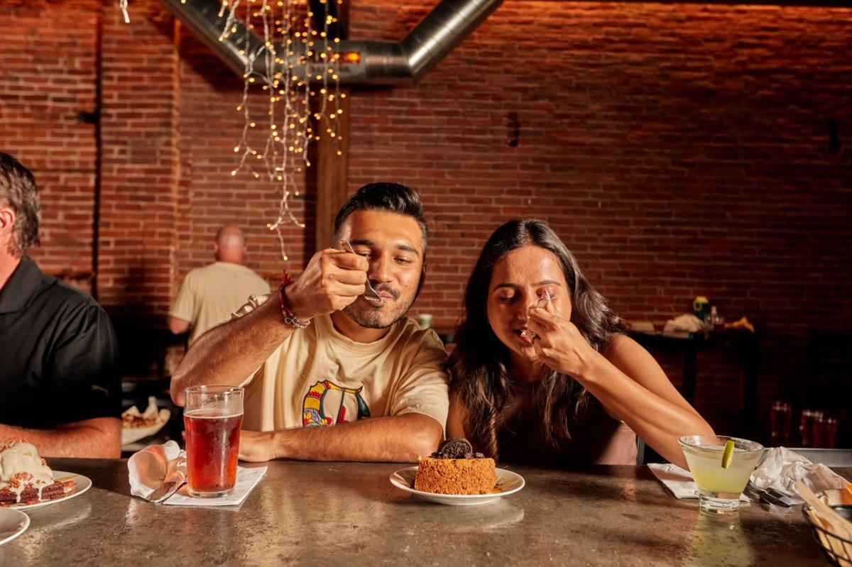 A couple sitting at a bar, eating a dessert. There is a brick wall behind them.