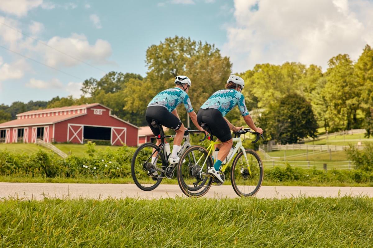Two people riding bikes in the summer, with a red barn behind them.