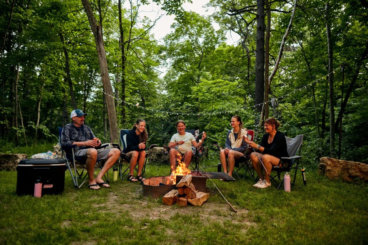 A family sitting around a campfire in the woods, holding skewers with marshmallows.