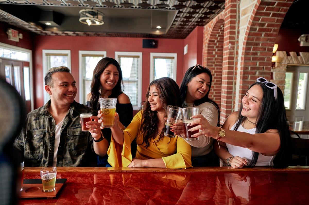 A group of friends holding drinks, going in for a "cheers". Three people are sitting at a bar and two people are standing behind them.