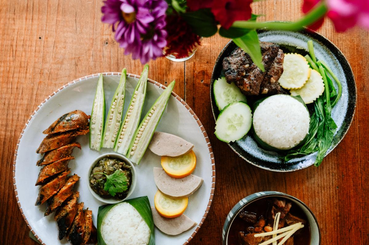An overhead view of a variety of Thai dishes served on a wooden table, including sliced spicy sausage, fresh okra, sticky rice wrapped in banana leaves, and grilled meats. A small vase of purple flowers sits at the top of the frame.