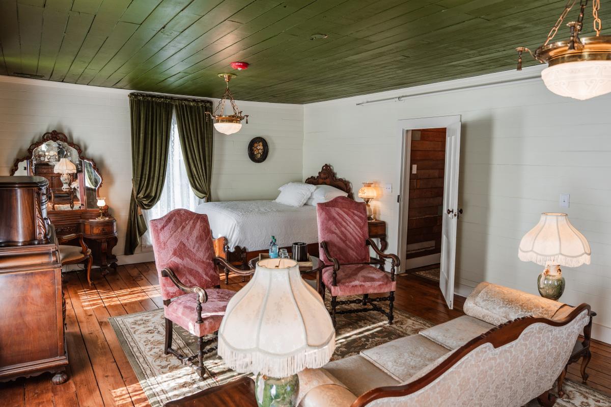 A historic bedroom at the Jarrett House featuring a dark green wooden plank ceiling, white plank walls, and wide-plank hardwood floors. The room is furnished with Victorian-era decor, including two pink velvet armchairs, a white bed, and a large antique wooden vanity. Sunlight streams across a patterned area rug, and several vintage lamps with fringed shades provide a warm glow.