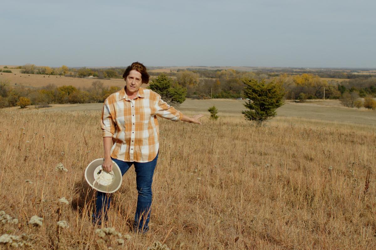 Woman standing in a field, holding her hat with one hand. With her other she points down a hill behind her.