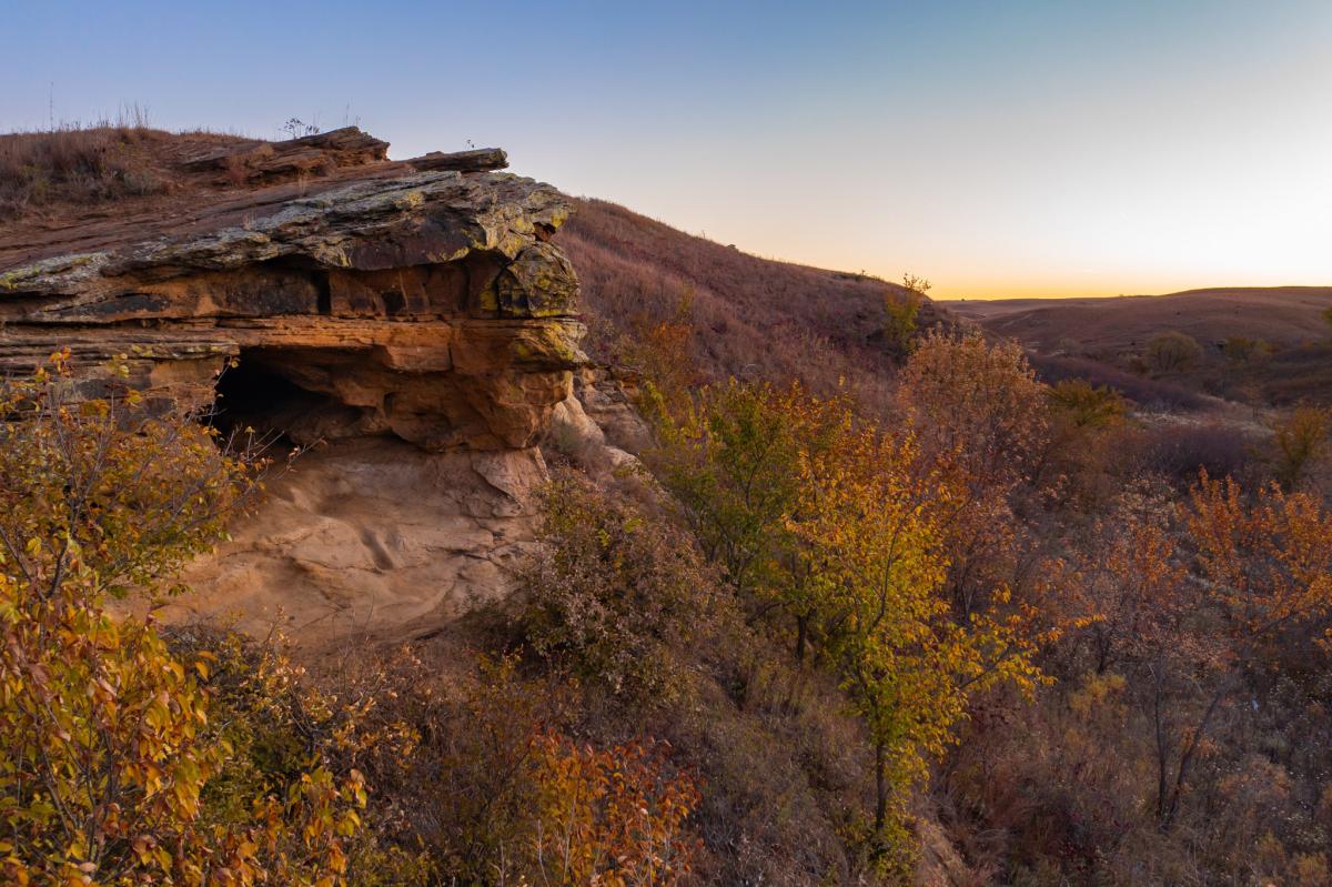 A straight on view of a rocky cave, surrounded by an autumn landscape.