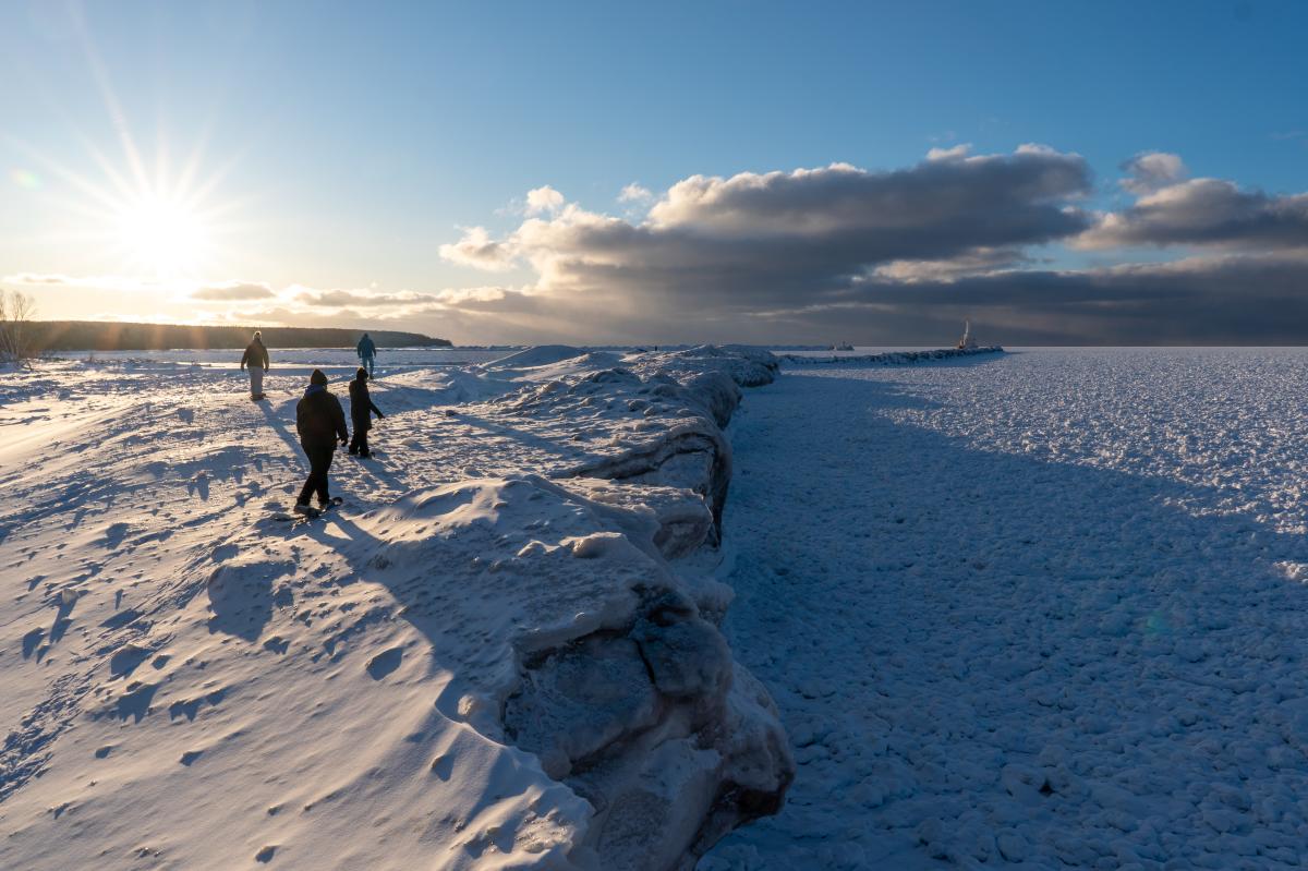 Two snowshoers trek along the frozen shoreline at Mclain State Park on a sunny day. Beautiful sun rays can be seen, as well as the Keweenaw North Entrance Light.