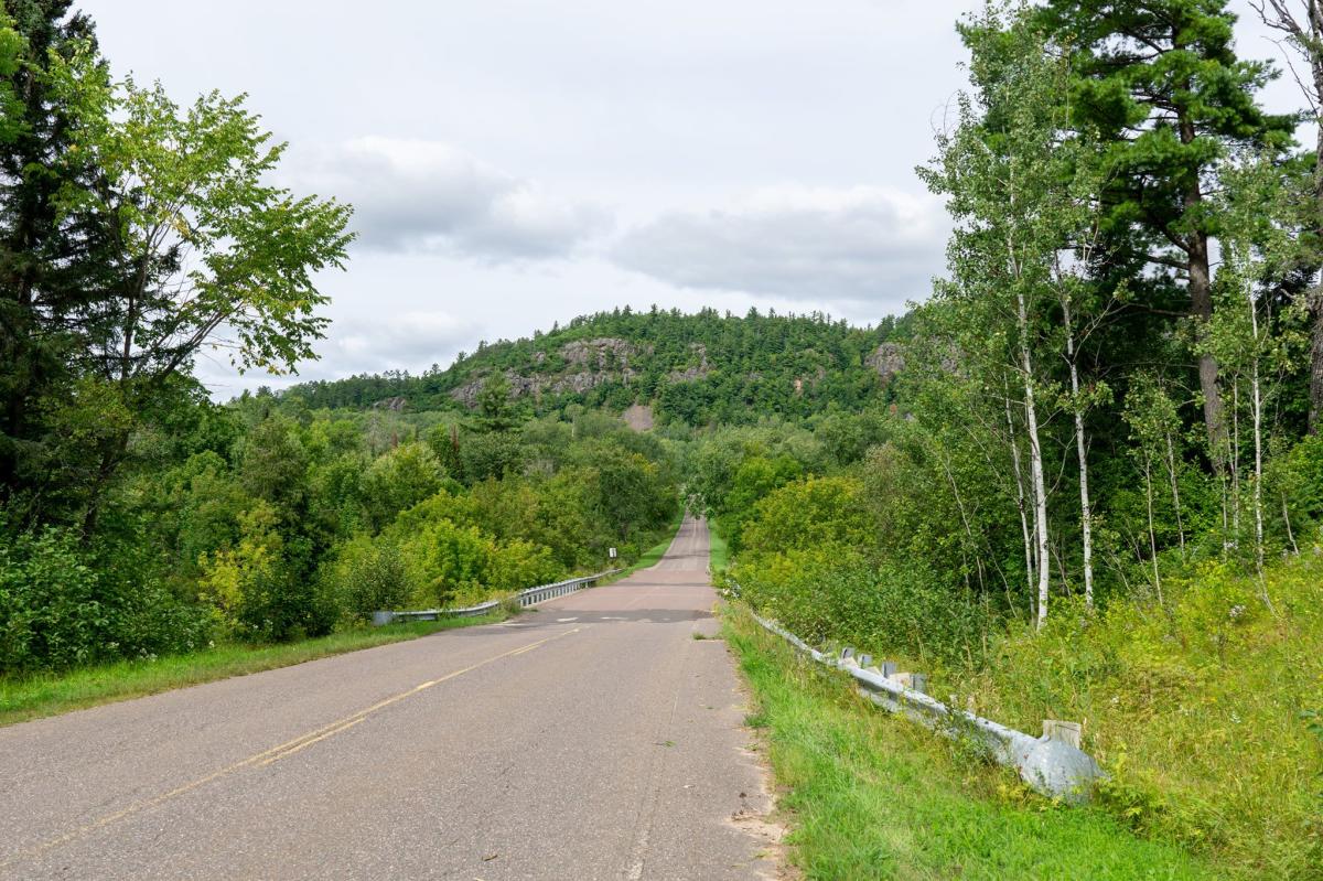 a quiet rural paved road stretches into the distance, a forested cliff on the horizon