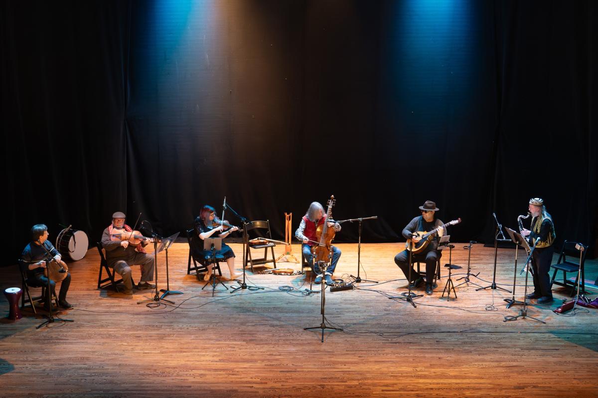 Photo shows Gerald Trimble and Jambaroque, a six-piece musical group sitting in a line and playing their instruments on stage at Liberty Hall in Lawrence, Kansas.